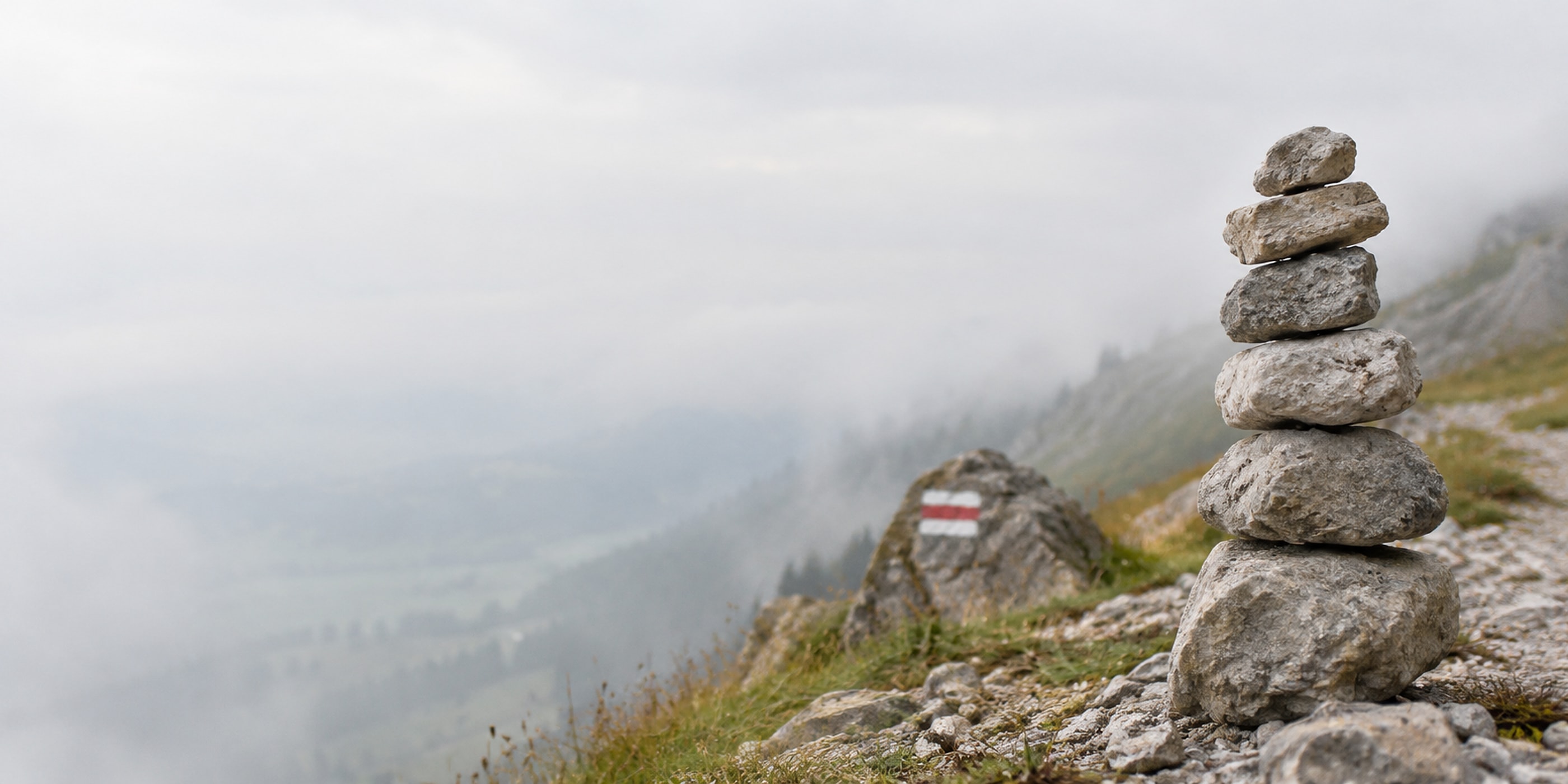 Un cairn au bord d’un sentier de montagne, près d’une balise blanc-rouge-blanc, au-dessus d’une vallée dans la brume.